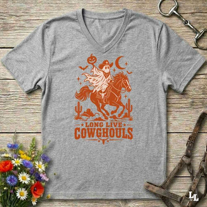 A grey V-neck t-shirt with a burnt orange graphic of a skeleton cowboy riding a horse through a desert scene with the text 'LONG LIVE COWGHOULS'. The shirt is displayed flat on a rustic wooden background with equestrian and floral props.