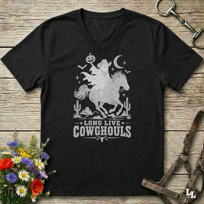 A black V-neck t-shirt with a burnt orange graphic of a skeleton cowboy riding a horse through a desert scene with the text 'LONG LIVE COWGHOULS'. The shirt is displayed flat on a rustic wooden background with equestrian and floral props.