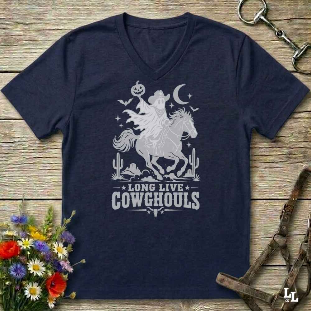 A navy blue V-neck t-shirt with a burnt orange graphic of a skeleton cowboy riding a horse through a desert scene with the text 'LONG LIVE COWGHOULS'. The shirt is displayed flat on a rustic wooden background with equestrian and floral props.