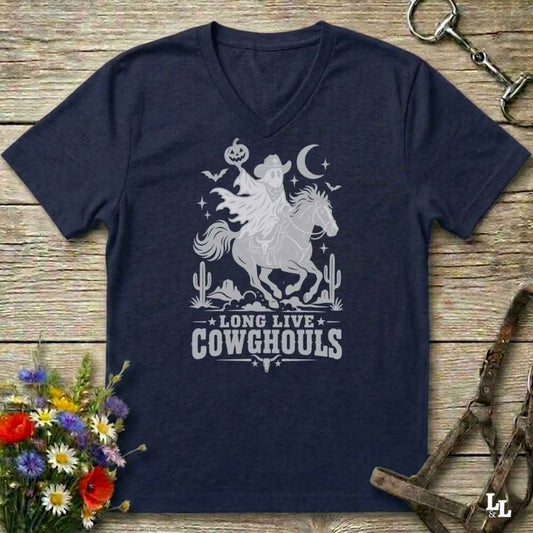 A navy blue V-neck t-shirt with a burnt orange graphic of a skeleton cowboy riding a horse through a desert scene with the text 'LONG LIVE COWGHOULS'. The shirt is displayed flat on a rustic wooden background with equestrian and floral props.