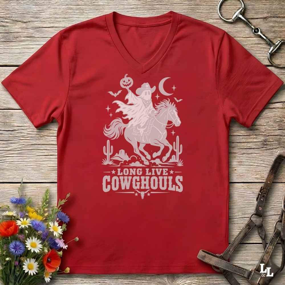 A red V-neck t-shirt with a burnt orange graphic of a skeleton cowboy riding a horse through a desert scene with the text 'LONG LIVE COWGHOULS'. The shirt is displayed flat on a rustic wooden background with equestrian and floral props.