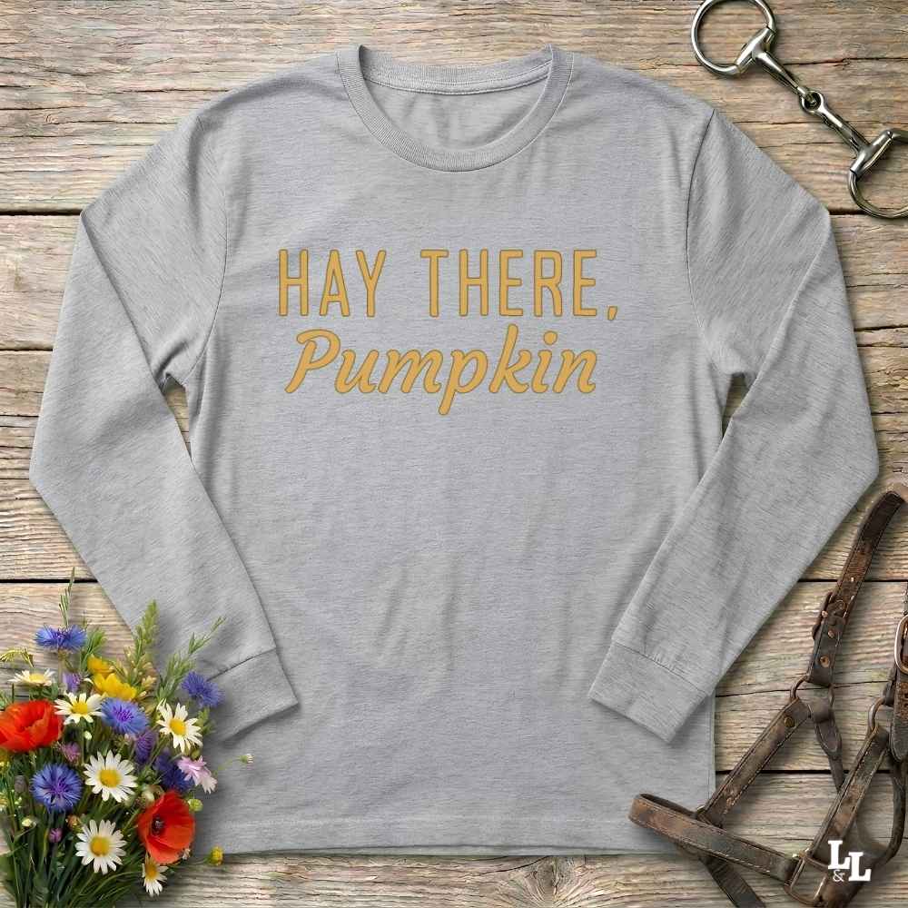 A light heather gray long-sleeve t-shirt with the phrase 'HAY THERE, Pumpkin' printed in gold across the chest, laid flat on a rustic wooden surface with decorative flowers and equestrian tack around it.