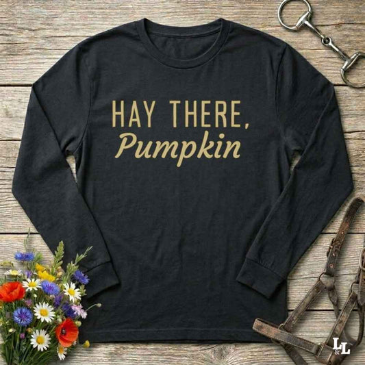A black long-sleeve t-shirt with the phrase 'HAY THERE, Pumpkin' printed in gold lettering, laid flat on a rustic wooden background with equestrian and floral props.