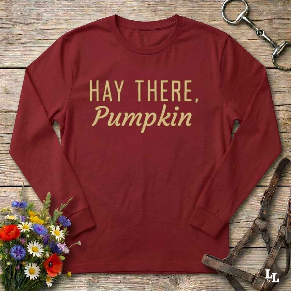 A long-sleeve crewneck shirt in deep burgundy with the text 'HAY THERE, Pumpkin' printed in gold across the chest, styled on a rustic wooden surface with equestrian tack and a small bouquet of flowers.