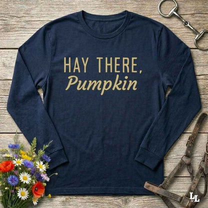 A navy blue long-sleeve t-shirt with the phrase 'HAY THERE, Pumpkin' printed in gold across the chest, styled in a flat-lay on a rustic wooden background with equestrian tack and a small flower bouquet.