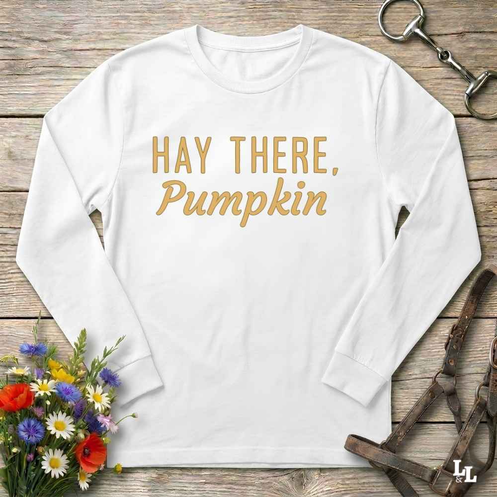 A white long-sleeve t-shirt with the phrase 'HAY THERE, Pumpkin' printed in gold lettering, laid flat on a rustic wooden background with decorative equestrian and floral props.
