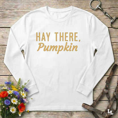 A white long-sleeve t-shirt with the phrase 'HAY THERE, Pumpkin' printed in gold lettering, laid flat on a rustic wooden background with decorative equestrian and floral props.