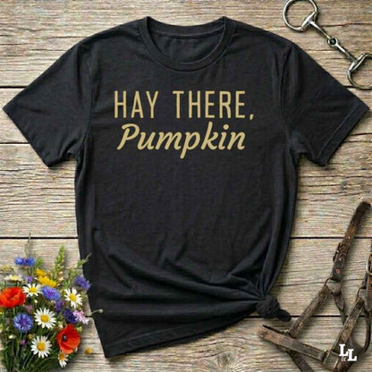 A black short-sleeve graphic T-shirt laid flat on a rustic wooden surface, with the phrase “HAY THERE, Pumpkin” printed in tan across the chest. The shirt is styled with flowers, a horse bit, and a leather halter around it.