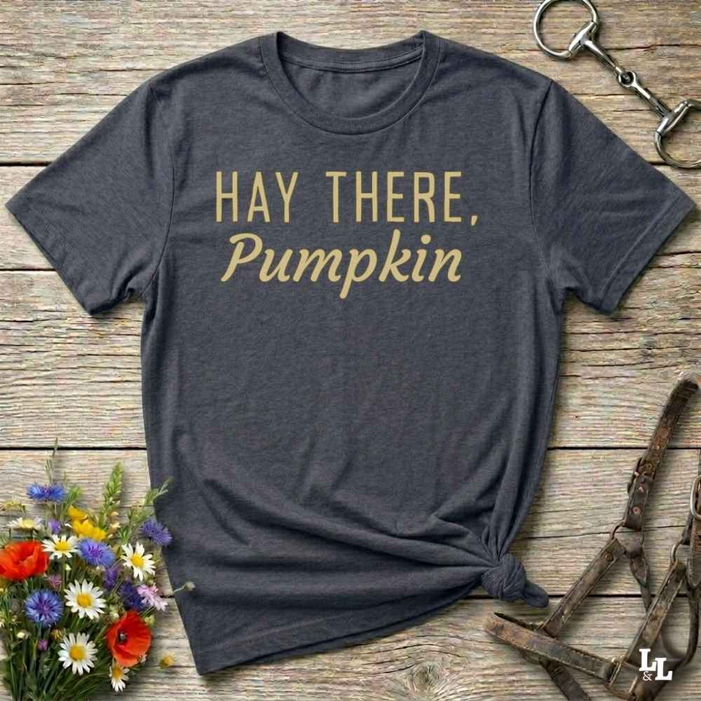A dark charcoal gray graphic t-shirt with the text 'HAY THERE, Pumpkin' printed in gold. The t-shirt is styled in a flat-lay arrangement on a rustic wooden background, accompanied by a small bouquet of wildflowers, an equestrian halter, and a metal tack clip.