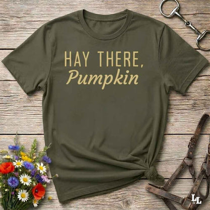An olive green t-shirt with the text 'HAY THERE, Pumpkin' printed in gold lettering, styled in a flat-lay on a rustic wooden background with equestrian-themed accessories and flowers.