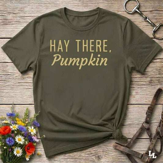 An olive green t-shirt with the text 'HAY THERE, Pumpkin' printed in gold lettering, styled in a flat-lay on a rustic wooden background with equestrian-themed accessories and flowers.