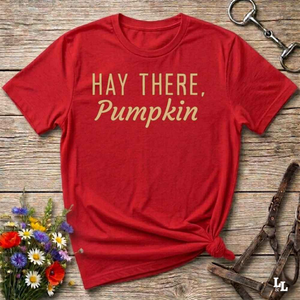 A red short-sleeve t-shirt with the text 'HAY THERE, Pumpkin' printed in a gold serif font, styled flat on a rustic wooden background with decorative flowers and horse tack.