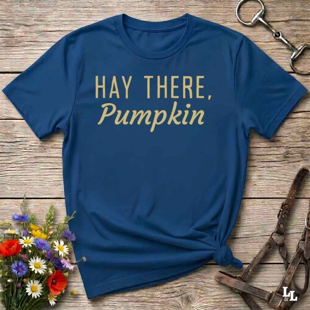 A navy blue short-sleeve t-shirt with the phrase 'HAY THERE, Pumpkin' printed in gold lettering, styled in a flat-lay on a rustic wooden background with flowers and horse tack props.