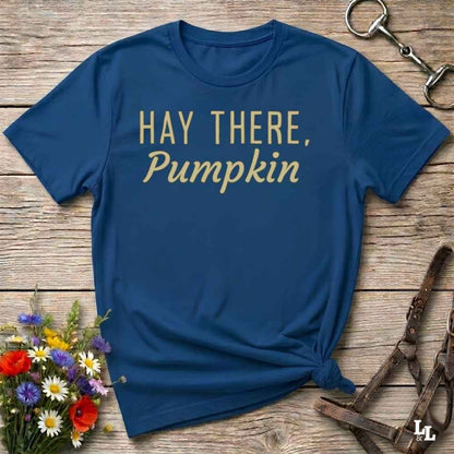 A navy blue short-sleeve t-shirt with the phrase 'HAY THERE, Pumpkin' printed in gold lettering, styled in a flat-lay on a rustic wooden background with flowers and horse tack props.