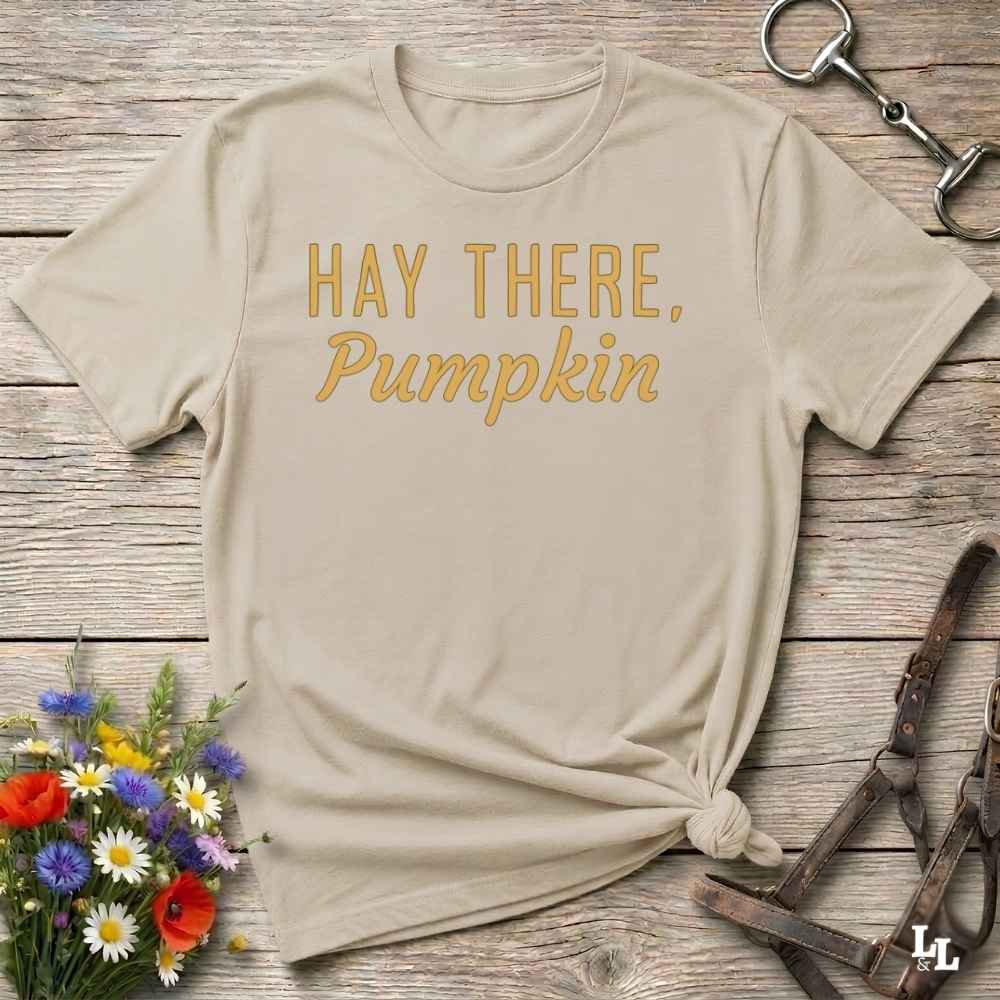 A beige graphic t-shirt with the text 'HAY THERE, Pumpkin' printed in gold on the front, styled in a flat-lay on a rustic wooden background with a horse bit, leather straps, and a small bouquet of wildflowers.