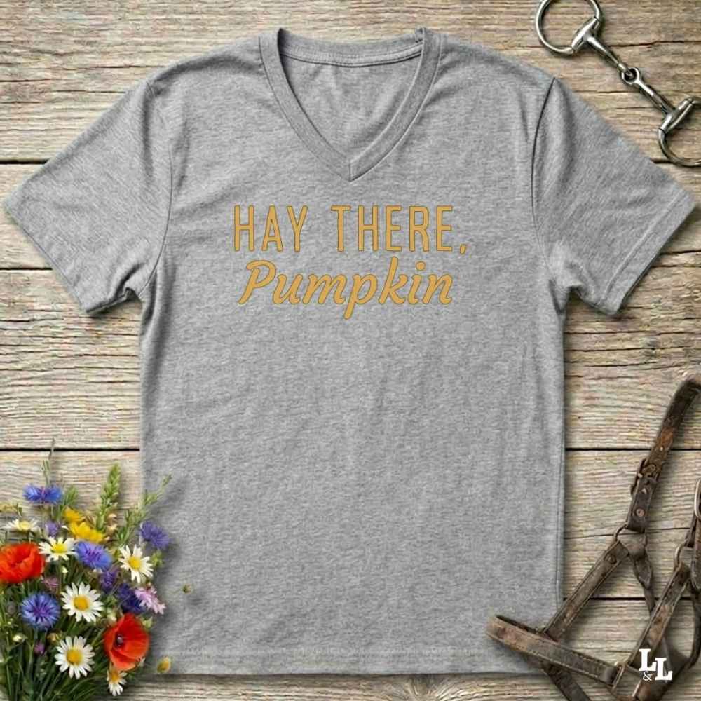 A heather gray V-neck t-shirt with the text 'HAY THERE, Pumpkin' printed in a golden-yellow font, laid flat on a rustic wooden background with equestrian and floral props.
