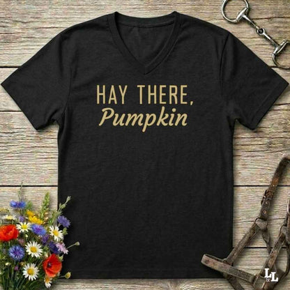 A black V-neck t-shirt with the phrase 'HAY THERE, Pumpkin' printed in gold lettering, displayed on a rustic wooden background with equestrian-themed props like a horse bit, bridle, and wildflowers.