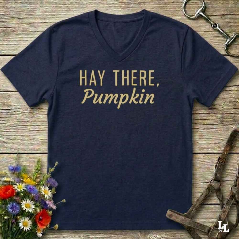 A navy blue V-neck t-shirt with the phrase 'HAY THERE, Pumpkin' printed in gold lettering across the chest, presented in a flat-lay arrangement on a rustic wooden background with equestrian tack and wildflowers.