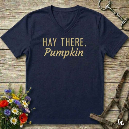 A navy blue V-neck t-shirt with the phrase 'HAY THERE, Pumpkin' printed in gold lettering across the chest, presented in a flat-lay arrangement on a rustic wooden background with equestrian tack and wildflowers.