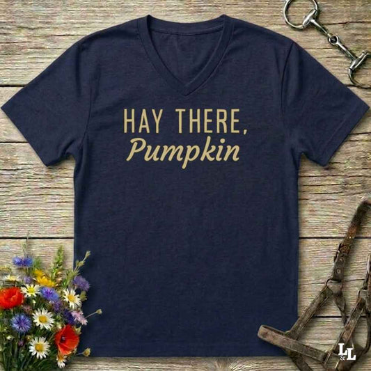 A navy blue V-neck t-shirt with the phrase 'HAY THERE, Pumpkin' printed in gold lettering across the chest, presented in a flat-lay arrangement on a rustic wooden background with equestrian tack and wildflowers.
