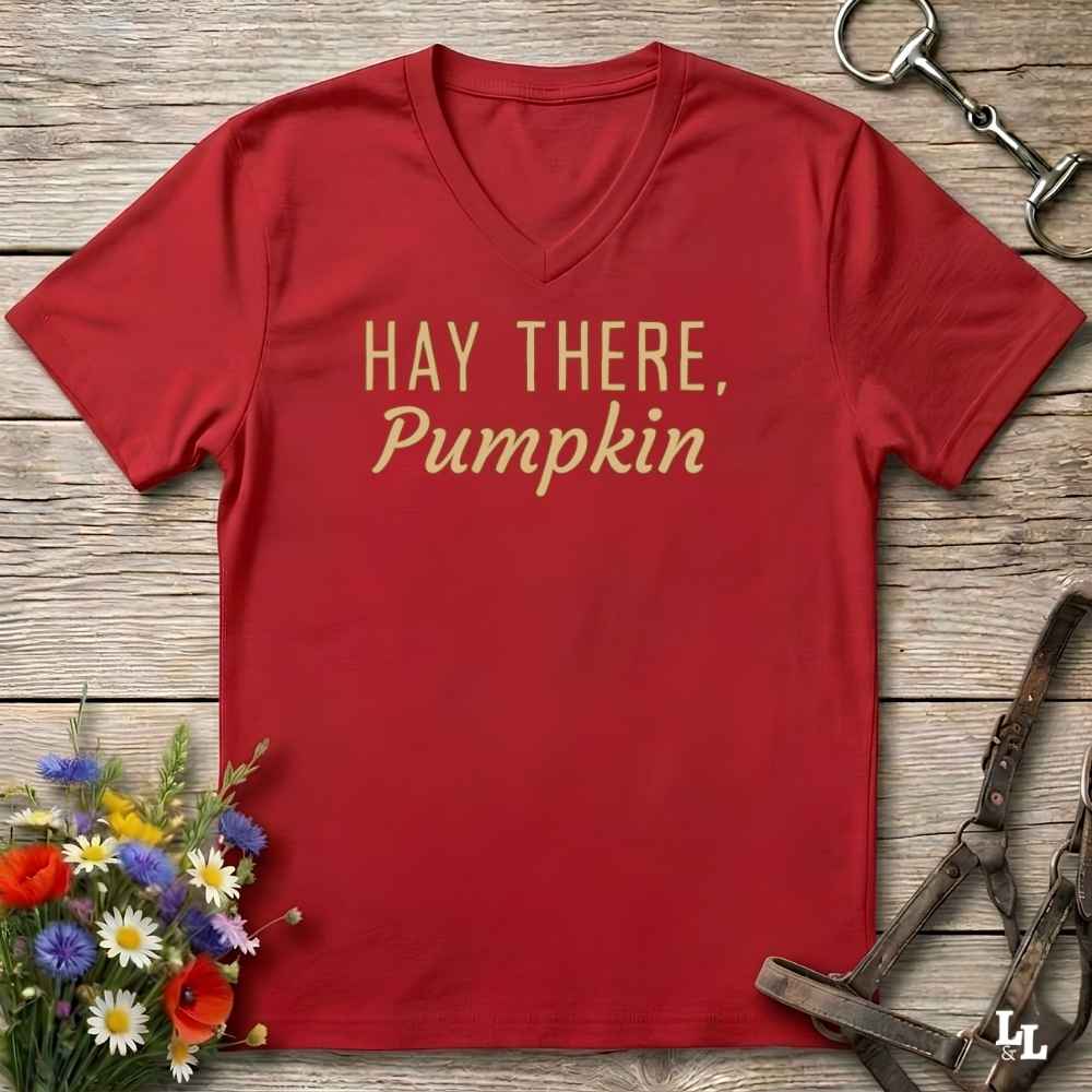 A red V-neck t-shirt with the text 'HAY THERE, Pumpkin' printed in gold on the front, styled in a flat-lay on a rustic wooden background with equestrian tack and wildflowers.