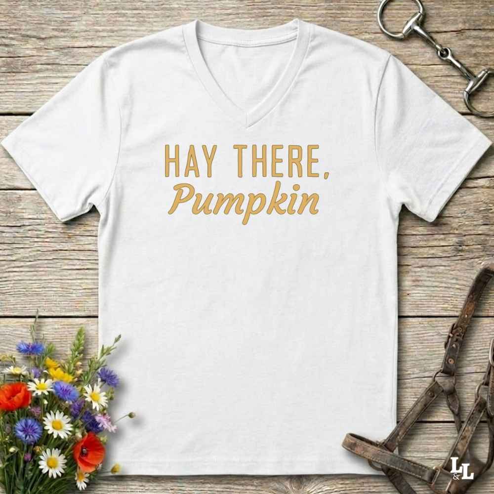 A white V-neck t-shirt with the text 'HAY THERE, Pumpkin' printed in a gold cursive font, displayed on a rustic wooden background with equestrian and floral props.