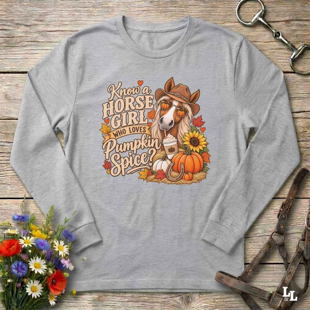 A light gray long-sleeve t-shirt with a fall-themed graphic print featuring a horse, pumpkins, sunflowers, and autumn leaves with the text 'Know a HORSE GIRL WHO LOVES Pumpkin Spice?'. The shirt is displayed flat on a rustic wooden background with decorative equestrian and floral props.