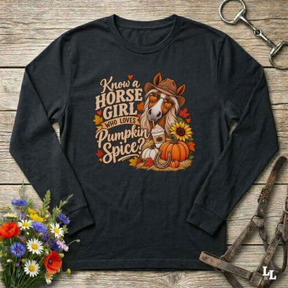 A black long-sleeve t-shirt with a colorful graphic of a horse in a cowboy hat surrounded by pumpkins, sunflowers, and autumn leaves, with the text 'Know a HORSE GIRL WHO LOVES Pumpkin Spice?'. The shirt is displayed flat on a rustic wooden background with decorative elements including a bouquet of wildflowers, a leather bridle, and a metal horse tack clip.