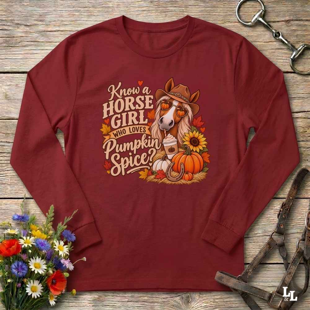 A long-sleeve crewneck shirt in burgundy red, laid flat on a rustic wooden background. The shirt features a detailed graphic on the front with a horse wearing a cowboy hat and sunglasses, surrounded by pumpkins, autumn leaves, a horseshoe, and text that reads 'Know a HORSE GIRL WHO LOVES Pumpkin Spice'.