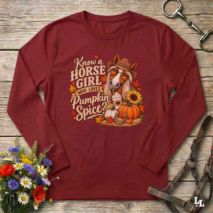 A long-sleeve crewneck shirt in burgundy red, laid flat on a rustic wooden background. The shirt features a detailed graphic on the front with a horse wearing a cowboy hat and sunglasses, surrounded by pumpkins, autumn leaves, a horseshoe, and text that reads 'Know a HORSE GIRL WHO LOVES Pumpkin Spice'.