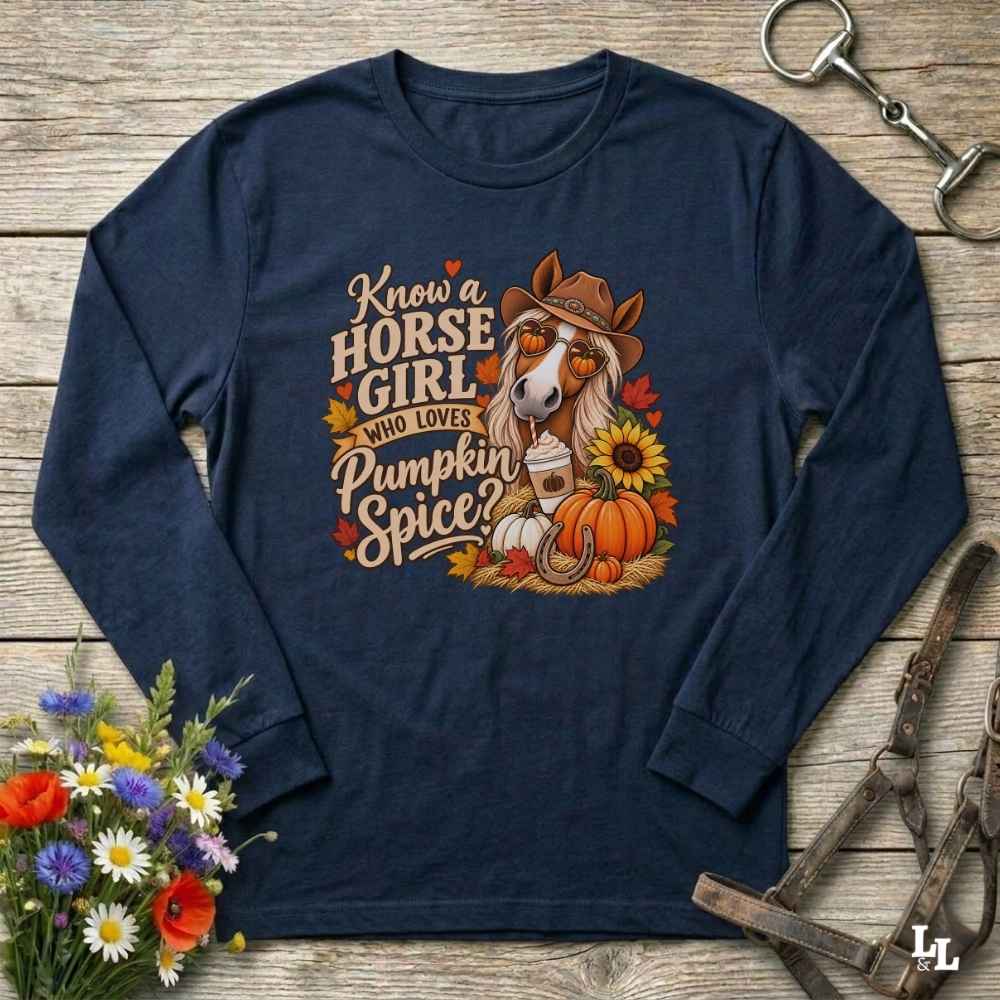 A navy blue long-sleeve t-shirt with a fall-themed graphic print, laid flat on a rustic wooden background with decorative props including a bouquet of flowers, horse tack, and a bridle. The graphic features a horse in a cowboy hat with the text 'Know a HORSE GIRL WHO LOVES Pumpkin Spice'.