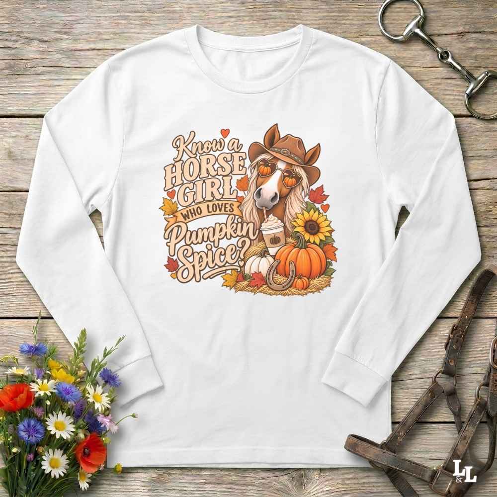 A white long-sleeve t-shirt with a colorful autumn-themed graphic print featuring a horse, pumpkins, sunflowers, and the text 'Know a HORSE GIRL WHO LOVES Pumpkin Spice?'. The shirt is displayed in a flat-lay arrangement on a rustic wooden background, styled with a bouquet of wildflowers, a leather bridle, and a horse bit.