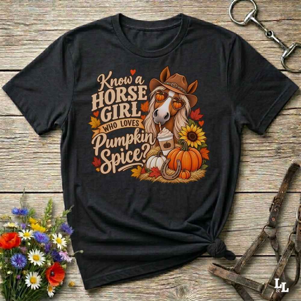 A black graphic t-shirt laid flat on a rustic wooden surface, featuring a fall-themed design with a horse wearing a cowboy hat, pumpkins, sunflowers, and autumn leaves. The text reads 'Know a HORSE GIRL WHO LOVES Pumpkin Spice'.