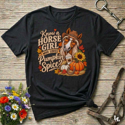 A black graphic t-shirt laid flat on a rustic wooden surface, featuring a fall-themed design with a horse wearing a cowboy hat, pumpkins, sunflowers, and autumn leaves. The text reads 'Know a HORSE GIRL WHO LOVES Pumpkin Spice'.