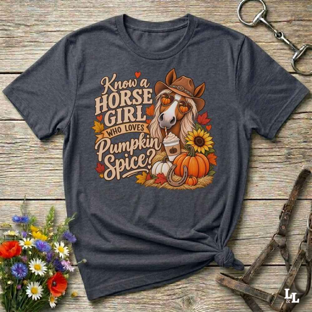 A dark heather gray graphic t-shirt laid flat on a rustic wooden surface, styled with a horse halter, flowers, and a small bundle of hay. The shirt features a colorful graphic of a horse wearing a cowboy hat with the text 'Know a HORSE GIRL WHO LOVES Pumpkin Spice'.