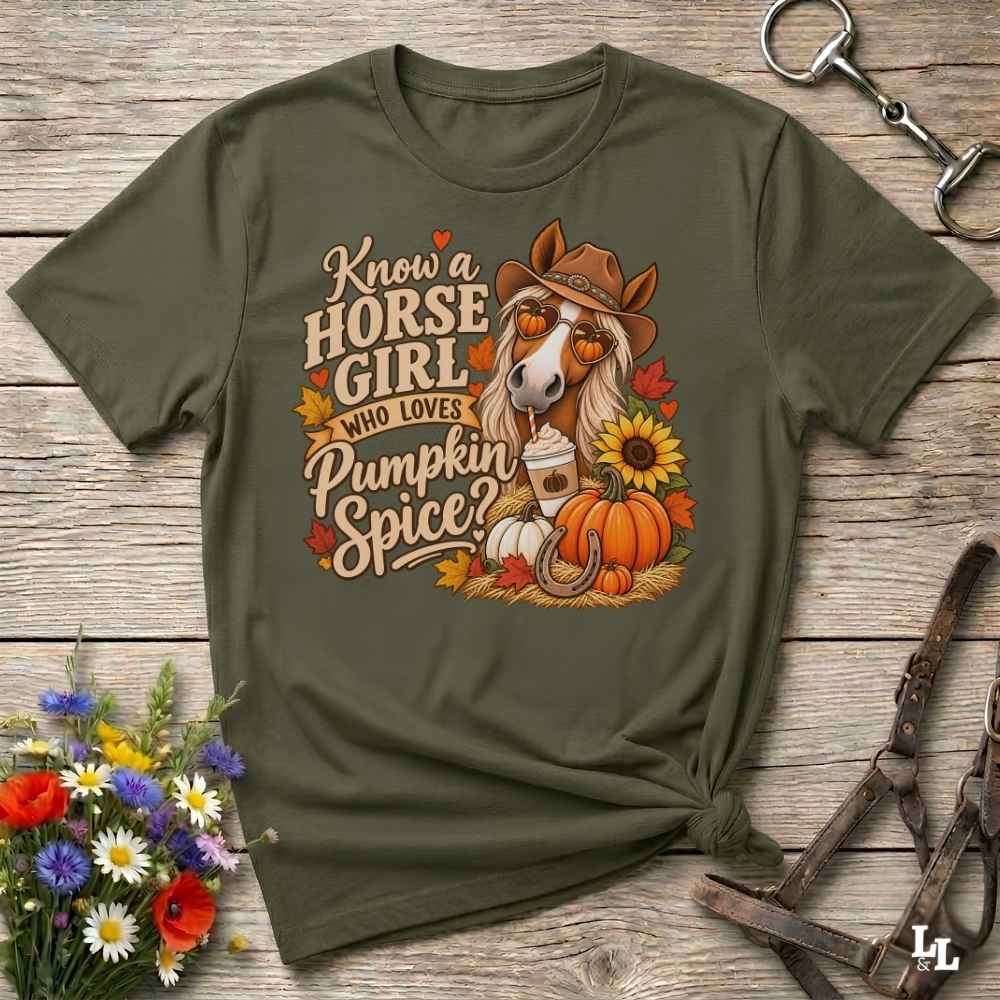 An olive green graphic t-shirt laid flat on a rustic wooden surface, styled with a horse halter, a bouquet of wildflowers, and a leather bridle. The t-shirt features a graphic of a horse wearing a cowboy hat and sunglasses, surrounded by pumpkins, fall leaves, and the text 'Know a HORSE GIRL WHO LOVES Pumpkin Spice'.