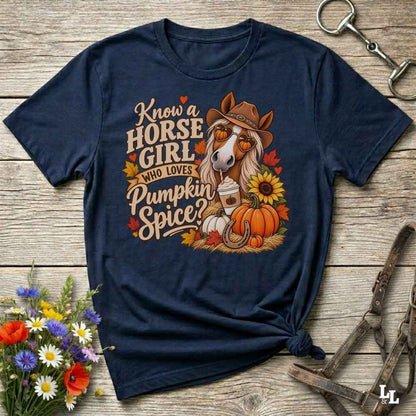 A navy blue graphic t-shirt laid flat on a rustic wooden surface, styled with a horse halter, flowers, and a small tie at the bottom right. The graphic features a cartoon horse wearing a cowboy hat and sunglasses, surrounded by pumpkins, autumn leaves, and the text 'Know a HORSE GIRL WHO LOVES Pumpkin Spice'.
