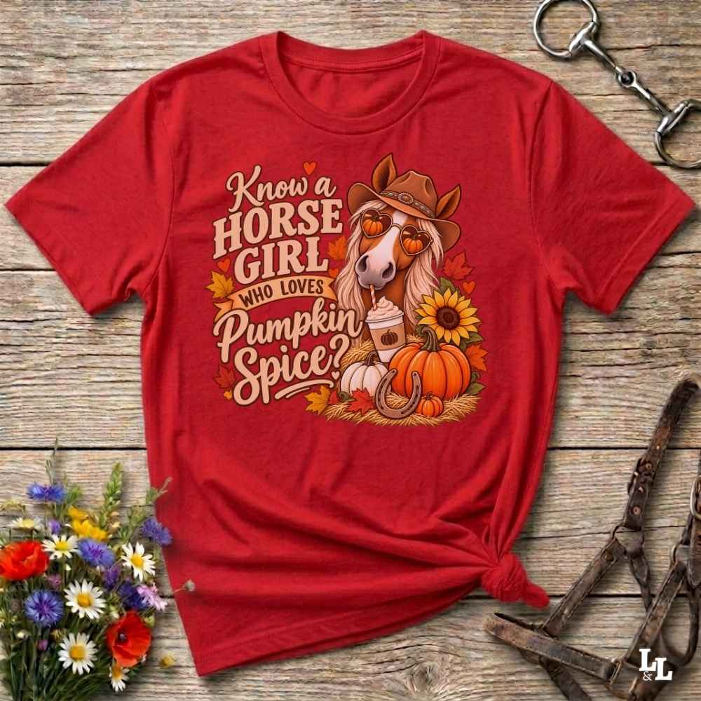 A red short-sleeve graphic T-shirt laid flat on a rustic wooden surface, styled with flowers, a horse bit, and a halter. The shirt features a large autumn-themed horse illustration and the text 'Know a HORSE GIRL WHO LOVES Pumpkin Spice'.
