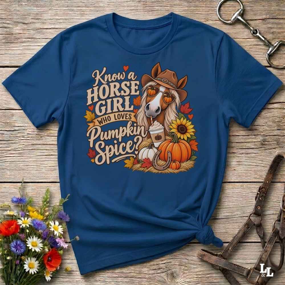 A navy blue graphic t-shirt laid flat on a rustic wooden surface, styled with a horse halter, a bouquet of wildflowers, and a metal tack piece. The t-shirt features a colorful graphic of a horse wearing a cowboy hat next to autumn-themed text that reads 'Know a HORSE GIRL WHO LOVES Pumpkin Spice?'.