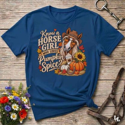 A navy blue graphic t-shirt laid flat on a rustic wooden surface, styled with a horse halter, a bouquet of wildflowers, and a metal tack piece. The t-shirt features a colorful graphic of a horse wearing a cowboy hat next to autumn-themed text that reads 'Know a HORSE GIRL WHO LOVES Pumpkin Spice?'.