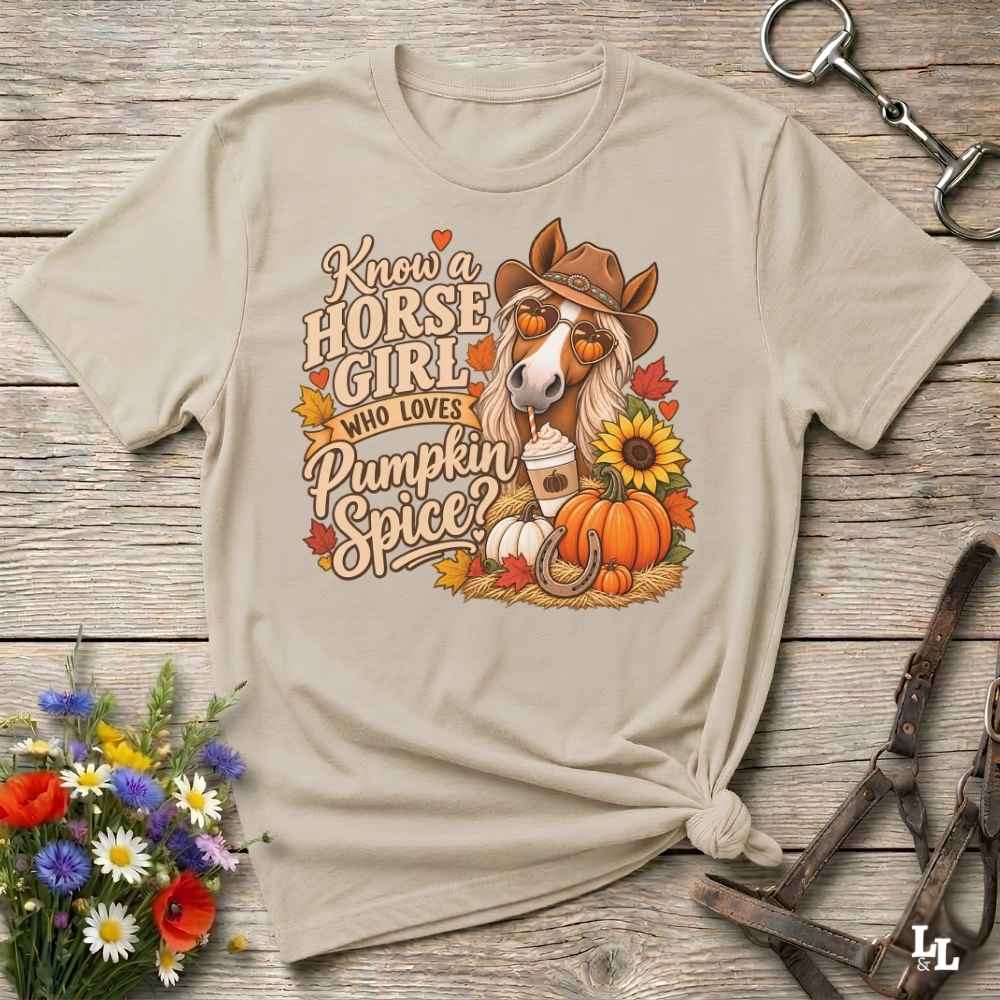 A beige graphic t-shirt featuring a fall-themed horse illustration with the text 'Know a HORSE GIRL WHO LOVES Pumpkin Spice?' surrounded by pumpkins, sunflowers, and autumn leaves, styled flat on a rustic wooden background with decorative flowers and leather tack.