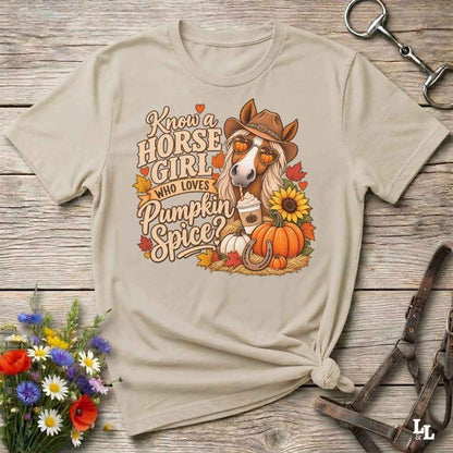 A beige graphic t-shirt featuring a fall-themed horse illustration with the text 'Know a HORSE GIRL WHO LOVES Pumpkin Spice?' surrounded by pumpkins, sunflowers, and autumn leaves, styled flat on a rustic wooden background with decorative flowers and leather tack.