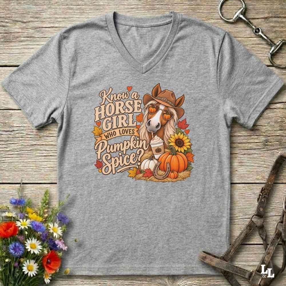 A heather gray V-neck t-shirt laid flat on a rustic wooden surface, featuring a colorful graphic of a horse in a cowboy hat with autumn-themed text reading 'Know a HORSE GIRL WHO LOVES Pumpkin Spice'. The shirt is styled with a bouquet of wildflowers, horse tack, and a leather strap.