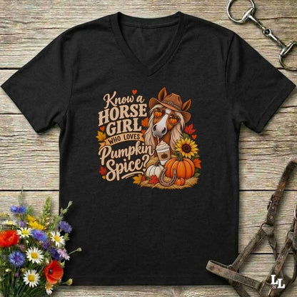 A black V-neck t-shirt laid flat on a rustic wooden surface, featuring a colorful graphic of a horse wearing a cowboy hat and sunglasses, surrounded by pumpkins, sunflowers, and the text 'Know a HORSE GIRL WHO LOVES Pumpkin Spice'. The shirt is styled with equestrian accessories and flowers.
