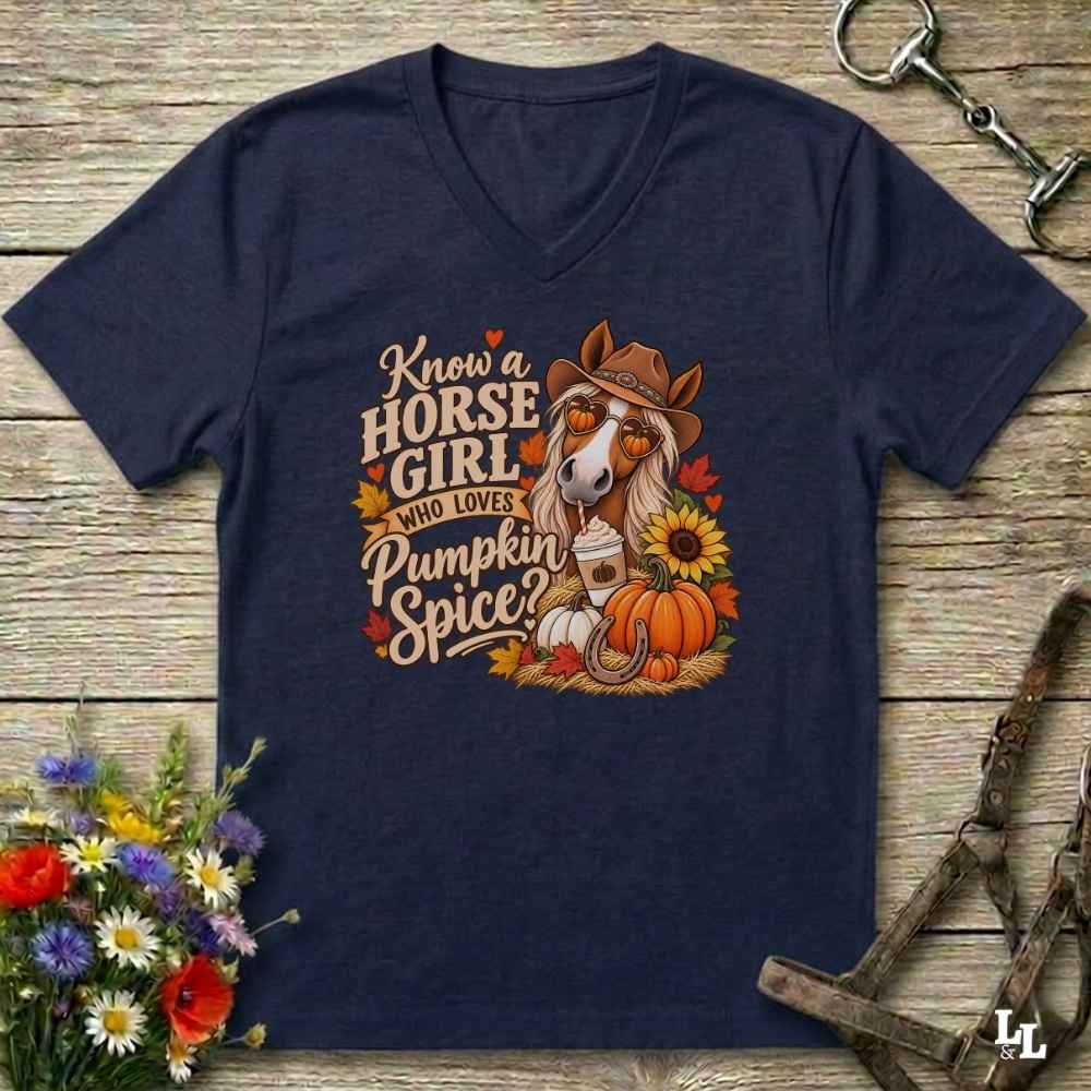 A navy blue V-neck t-shirt laid flat on a rustic wooden surface, featuring a colorful graphic of a horse wearing a cowboy hat with the text 'Know a HORSE GIRL who LOVES Pumpkin Spice'. The shirt is styled with equestrian tack, a bouquet of wildflowers, and autumn leaves.
