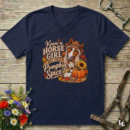 A navy blue V-neck t-shirt laid flat on a rustic wooden surface, featuring a colorful graphic of a horse wearing a cowboy hat with the text 'Know a HORSE GIRL who LOVES Pumpkin Spice'. The shirt is styled with equestrian tack, a bouquet of wildflowers, and autumn leaves.