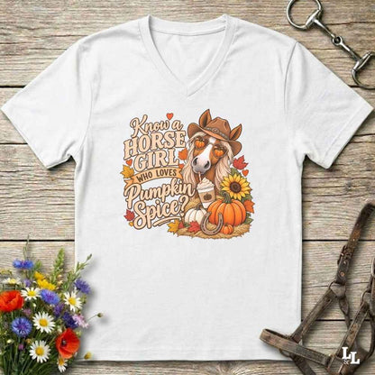 A white V-neck t-shirt laid flat on a rustic wooden surface, featuring a colorful graphic of a horse wearing a cowboy hat with the text 'Know a HORSE GIRL WHO LOVES Pumpkin Spice' and autumn-themed illustrations including pumpkins, sunflowers, and leaves. The shirt is styled with horse tack and flowers around it.