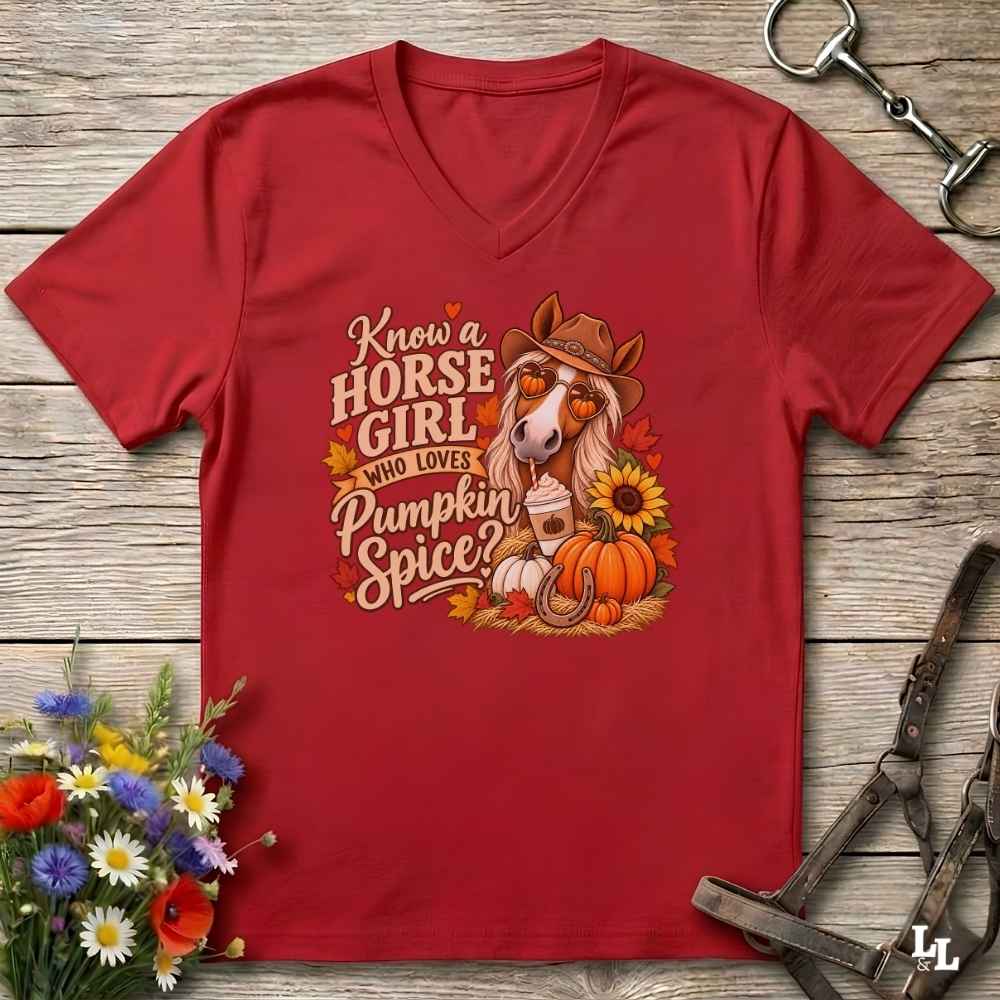 A red V-neck t-shirt laid flat on a rustic wood background, featuring a colorful graphic of a horse wearing a cowboy hat with the text 'Know a HORSE GIRL WHO LOVES Pumpkin Spice'. The shirt is styled with equestrian accessories (bridle and horsebit) and a small bouquet of wildflowers.