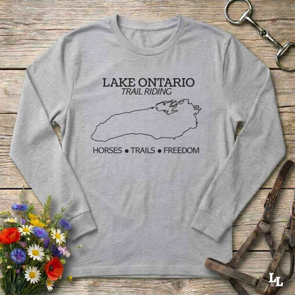 A light gray long-sleeve t-shirt with a black graphic print of Lake Ontario and the text 'LAKE ONTARIO TRAIL RIDING' and 'HORSES • TRAILS • FREEDOM'. The shirt is displayed flat on a rustic wooden background with equestrian accessories and a flower arrangement.