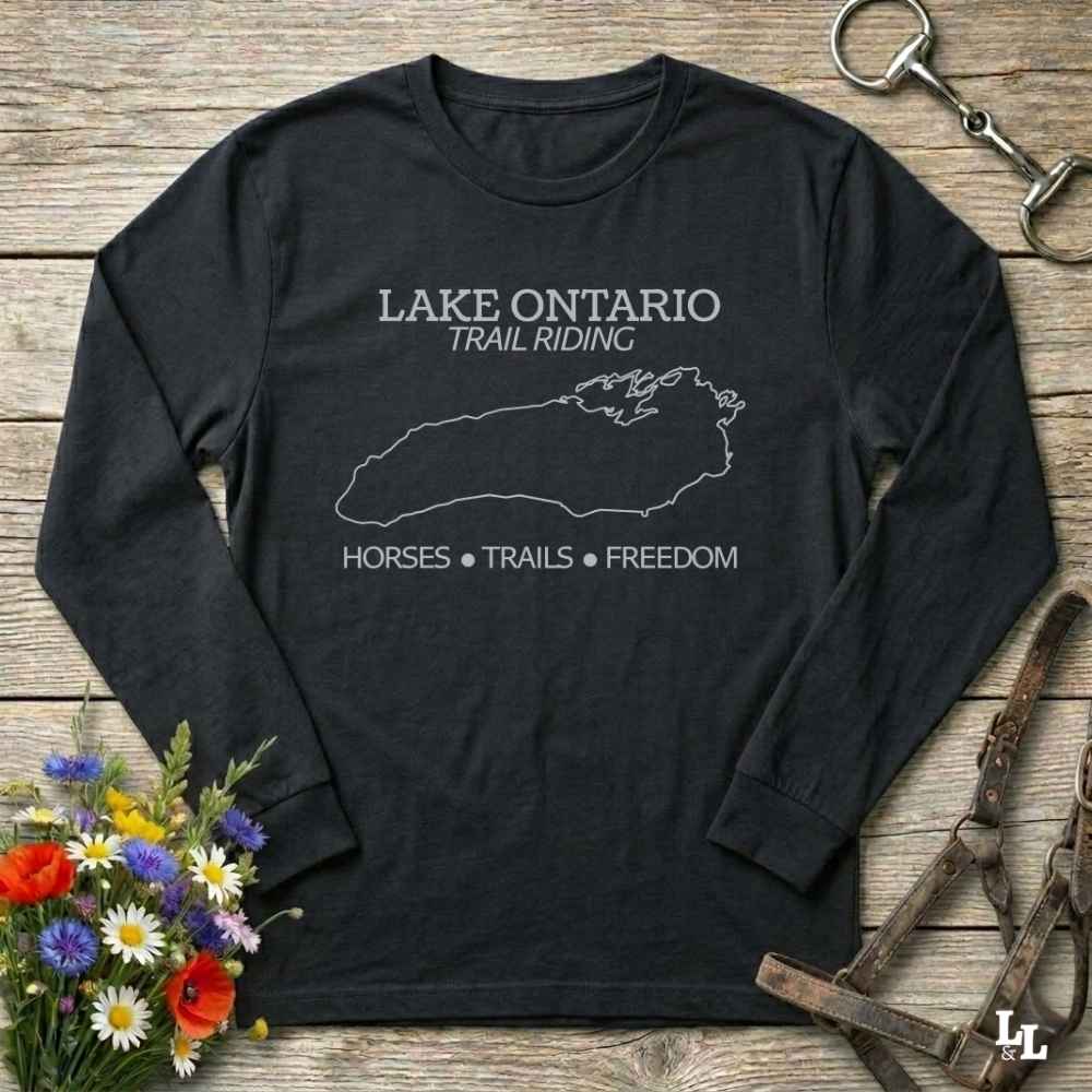 A black long-sleeve t-shirt with a white graphic print reading 'LAKE ONTARIO TRAIL RIDING' and 'HORSES • TRAILS • FREEDOM' with a lake outline, laid flat on a rustic wood background with flowers and horse tack props.